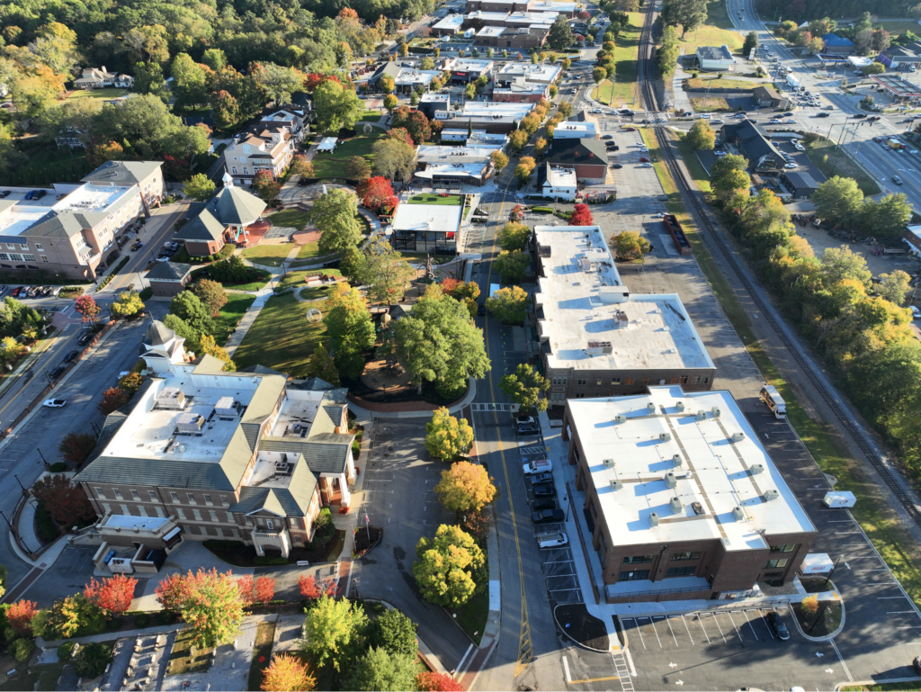 Aerial view of a small town with various buildings, roads, and trees. The image captures greenery and a few vehicles parked, with a mix of residential and commercial structures.